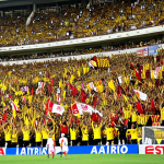 **

A vibrant, packed Estadio Monumental during a Universitario de Deportes vs. Alianza Lima "Clásico" match. Focus on the sea of yellow and red (Universitario's colors) in the stands, with fans chanting, waving flags, and the overall electric atmosphere. Include the stadium's vast size and architectural details. Capture the intensity and passion of the game.

**